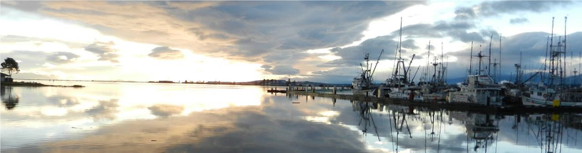 A photograph of the Comox Marina at sunrise. The image shows fishing boats reflected on the water, which also reflects the sun's glow and clouds.